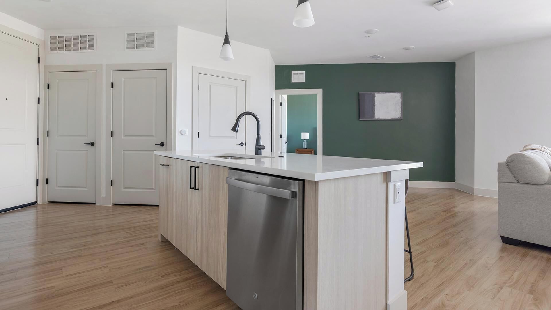 Una cocina moderna con lavavajillas, un lavamanos y una encimera. Hay dos puertas blancas, una pared verde y un sofá gris en la sala de estar.