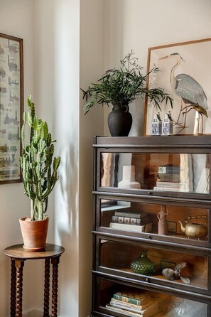 A cactus on a small table, a bookshelf with various items, and a framed picture of a bird on the wall. - 7BR Retreat, Walkers Paradise Near Washington Park (Cincinnati)