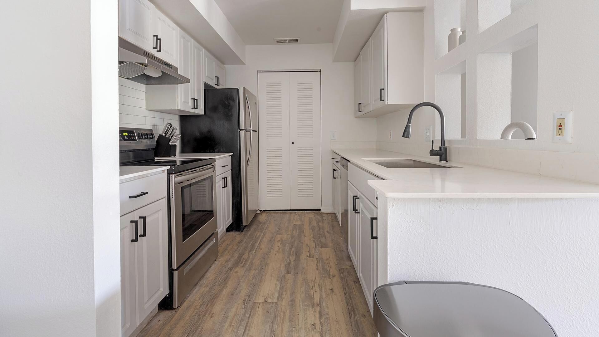 Cocina compacta con muebles blancos, refrigerador de acero inoxidable, horno empotrado y un lavamanos con grifo moderno.