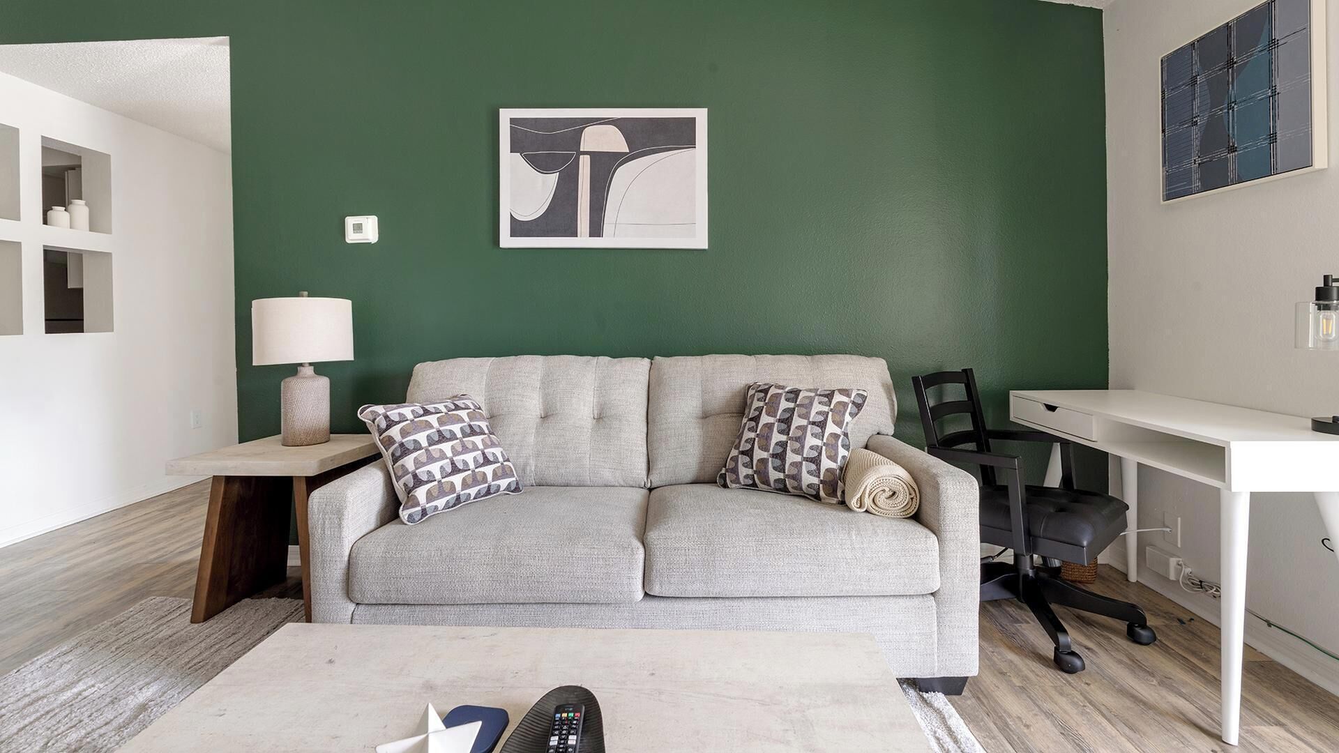 A living room with a grey sofa, patterned cushions, a small table with a lamp, a desk, and a chair.