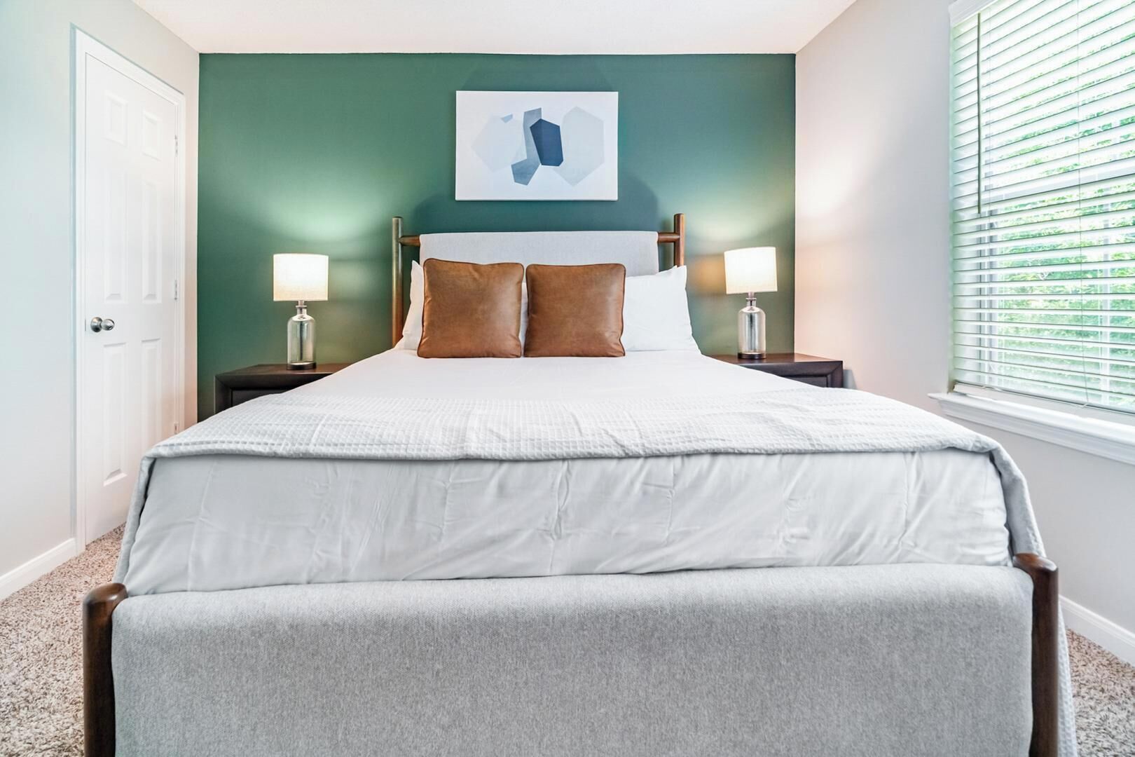 A neatly arranged bedroom with a bed, two bedside tables, a nightstand, a wall-mounted artwork, and a window with blinds.