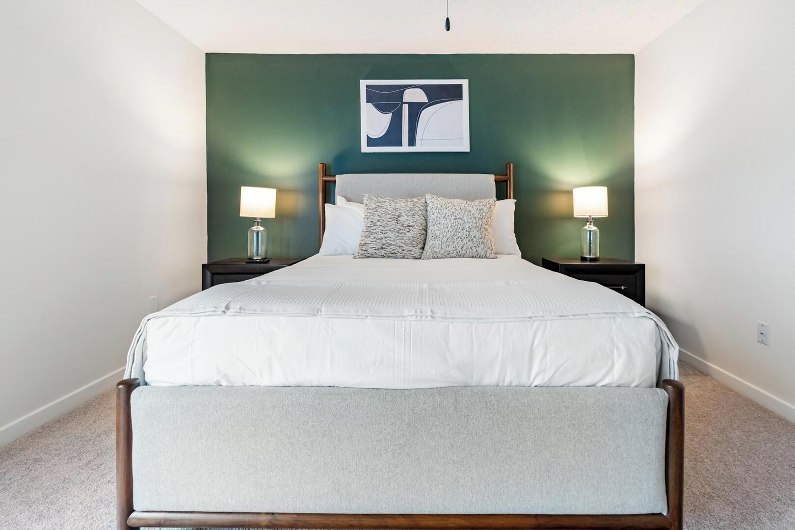 A large bed with a wooden frame, two bedside tables with lamps, a green accent wall, and a framed abstract artwork above the bed.