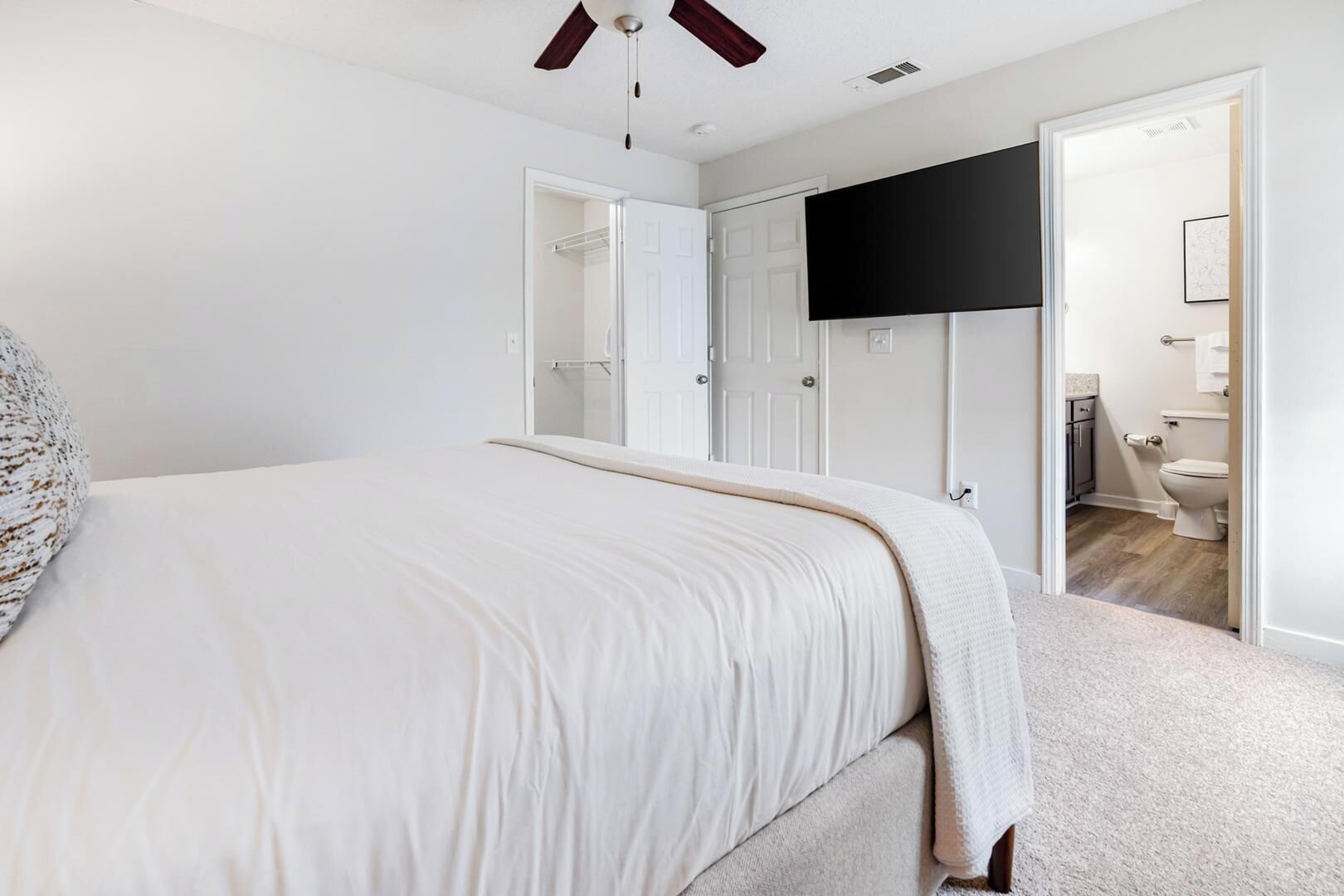 Une chambre à coucher avec un lit, un ventilateur de plafond, un téléviseur fixé au mur et une salle de bain visible à travers une porte ouverte.