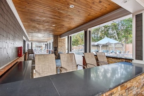 A covered outdoor seating area with a stone wall, wooden ceiling, and large windows overlooking a pool. - Landing | Stylish 3BD, Game Room, Clubhouse (Gulf Hills)