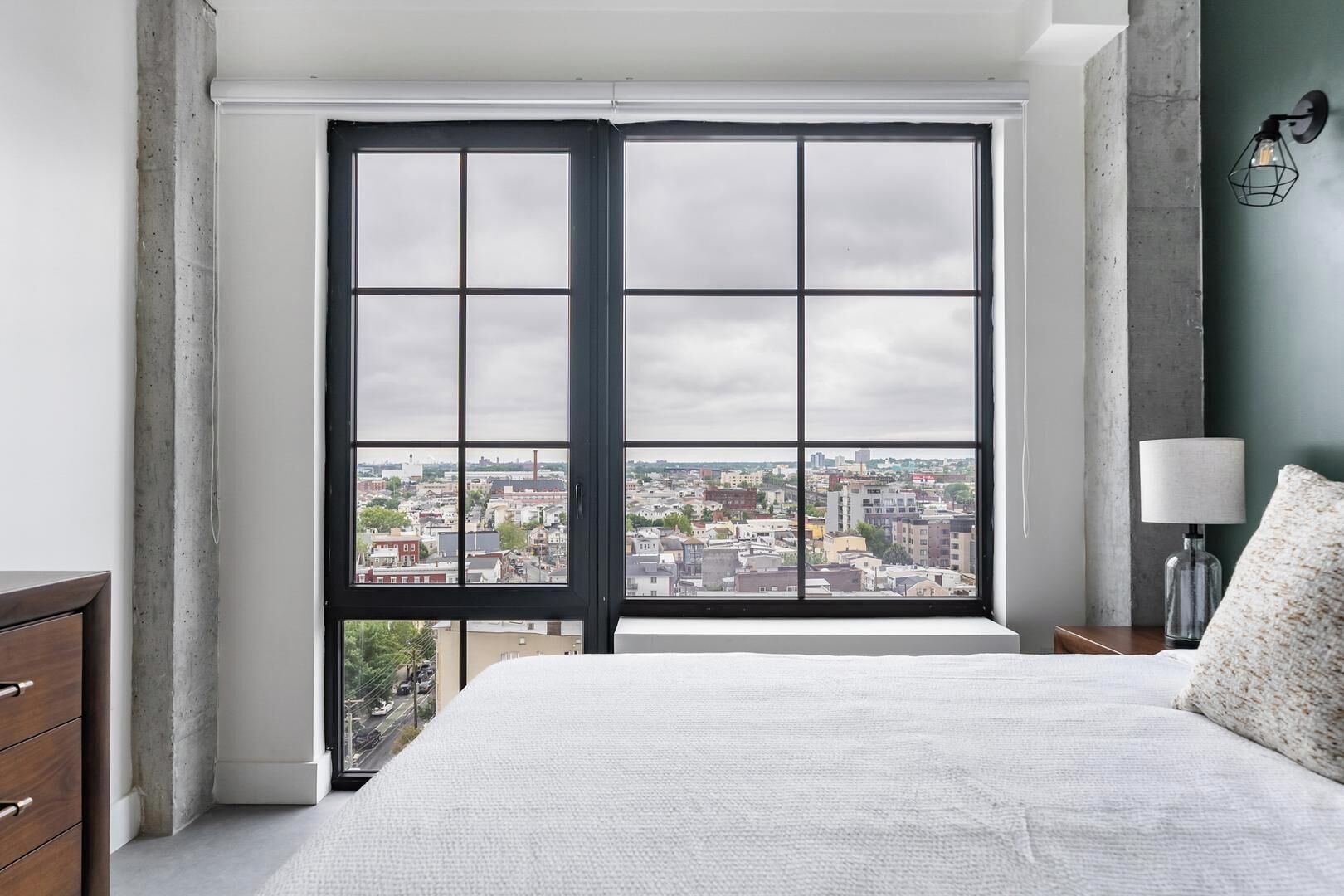 A bedroom with a large window, a bed, a nightstand, and a lamp.