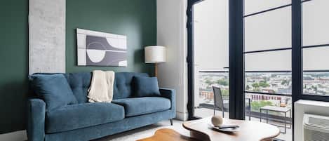 Een moderne woonkamer met een blauwe bank, een salontafel en uitzicht op een stadslandschap door grote ramen.