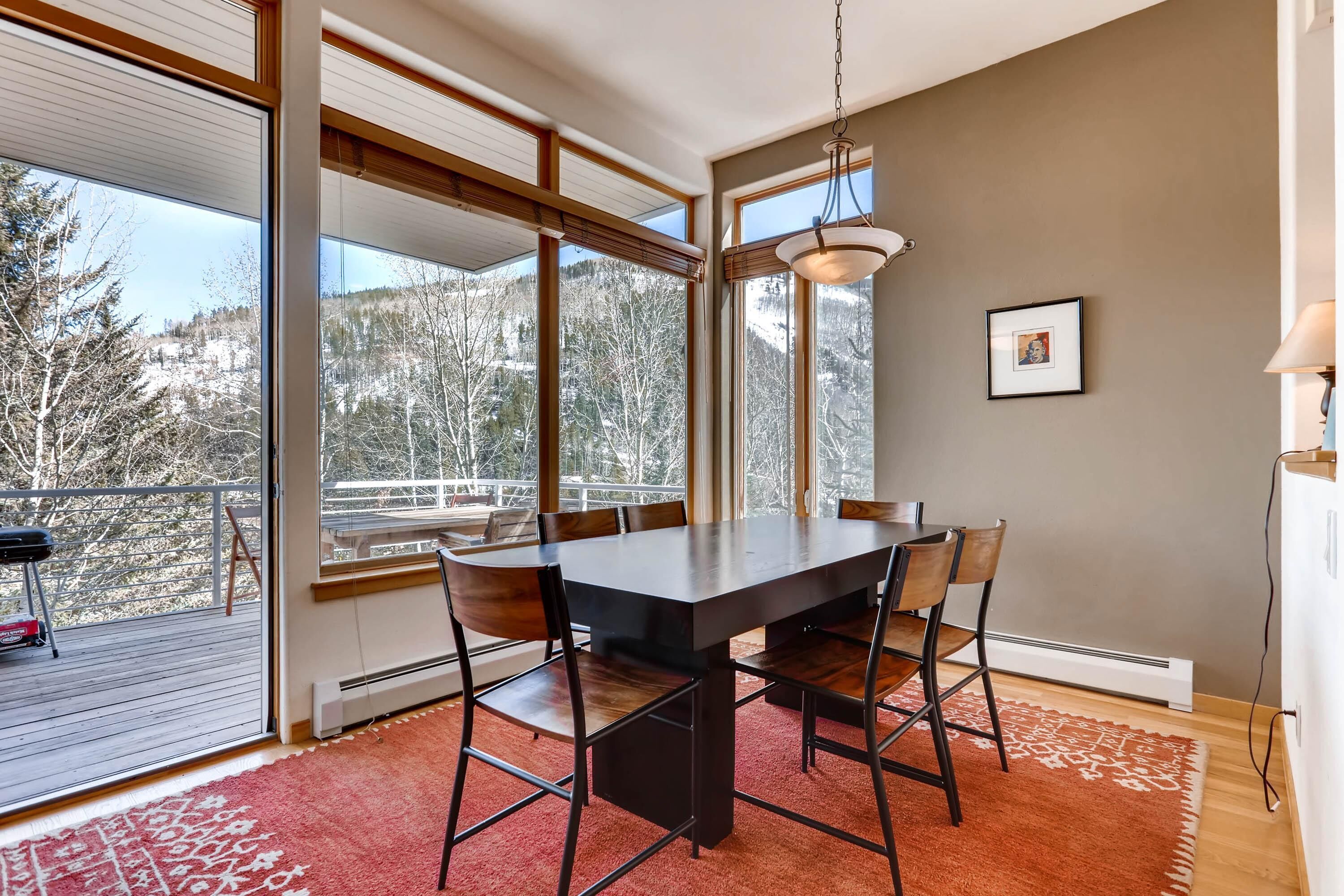 Un espace repas comprenant une grande table, des chaises et une vue sur les arbres, visible à travers de grandes fenêtres.