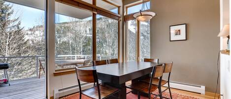 Un espace repas comprenant une grande table, des chaises et une vue sur les arbres, visible à travers de grandes fenêtres.