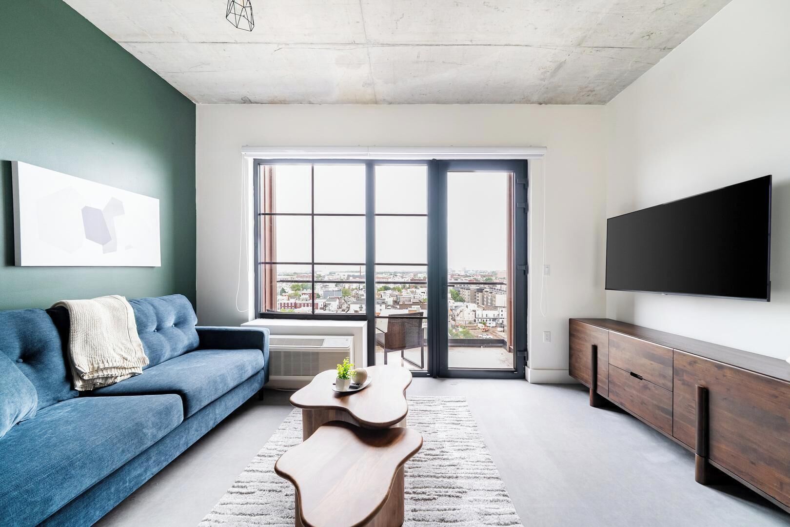 A modern living room with a blue sofa, a wooden coffee table, a TV on the wall, and a large window with a city view.