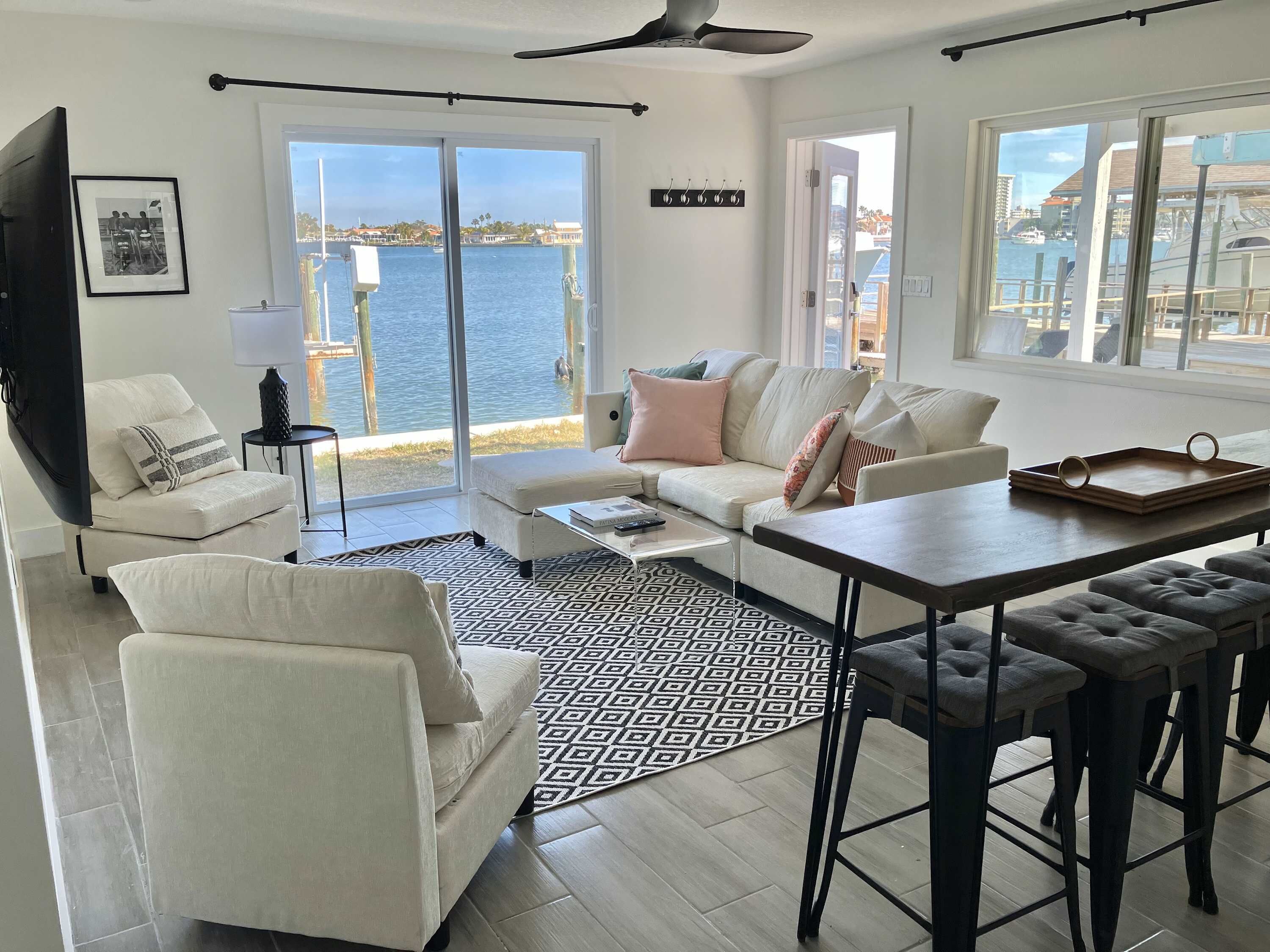 A modern living room with a sofa, coffee table, and bar stools. A large window offers a view of the water and boats.