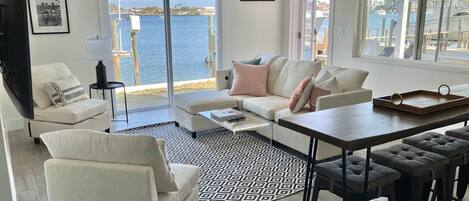 A modern living room with a sofa, coffee table, and bar stools. A large window offers a view of the water and boats.