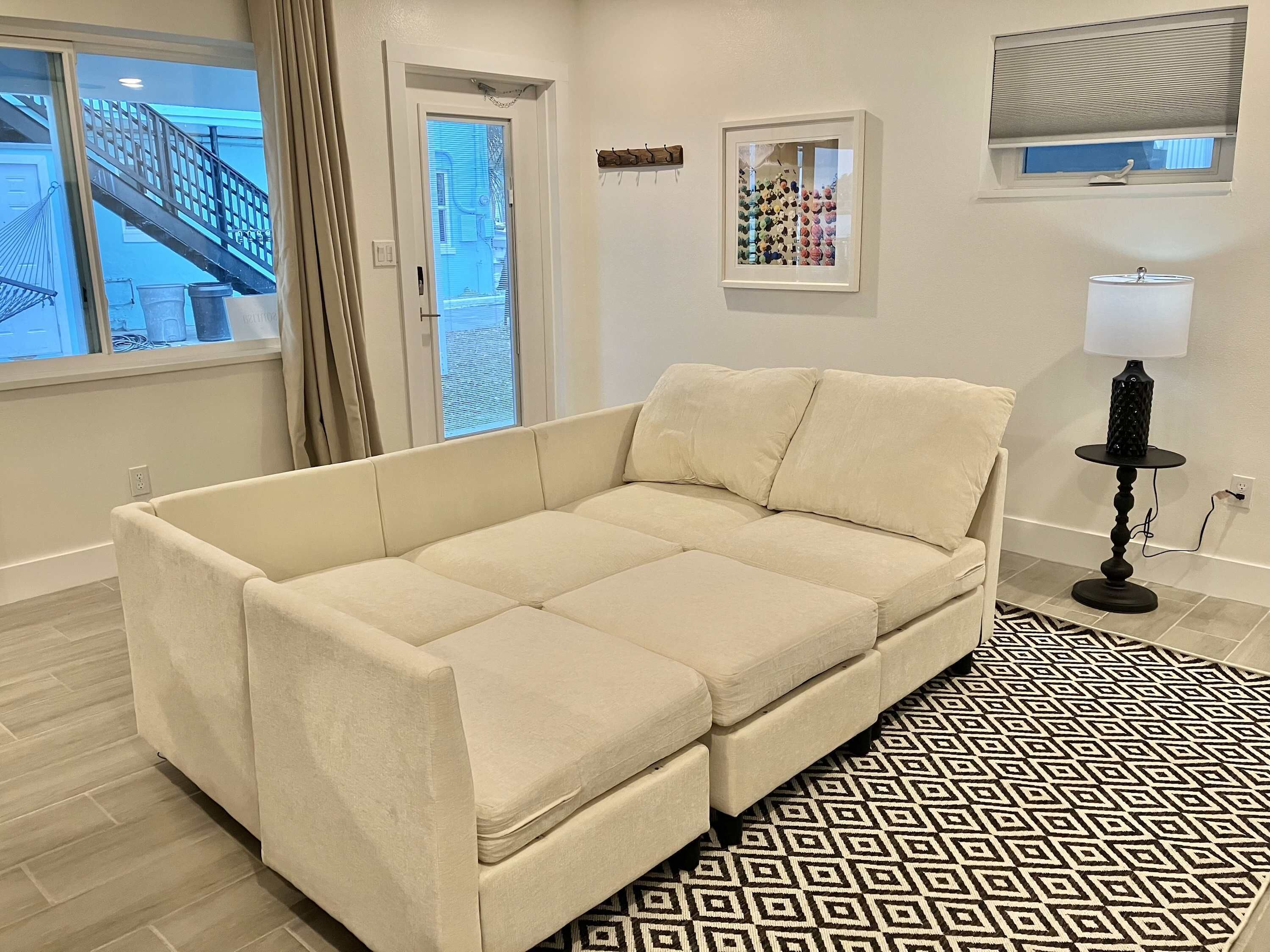 Un sofá color beige en una habitación con un tapete estampado, un cuadro en la pared y una ventana con persianas.