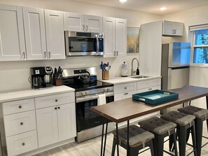 A modern kitchen with white cabinetry, stainless steel appliances, a coffee maker, and a bar stool area. - CWB Retreats at Bayside Suite 2 & View 3 (Clearwater)