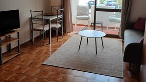 Un salon avec un téléviseur, une table à manger avec des chaises, un canapé, un divan rouge et une vue sur la rue à travers une porte vitrée.