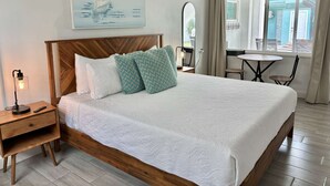 Een keurig opgemaakt hotelkamer met een houten hoofdeinde, een bed met witte lakens, een nachtkastje met een lamp, een klein tafeltje met stoelen en een raam met gordijnen.