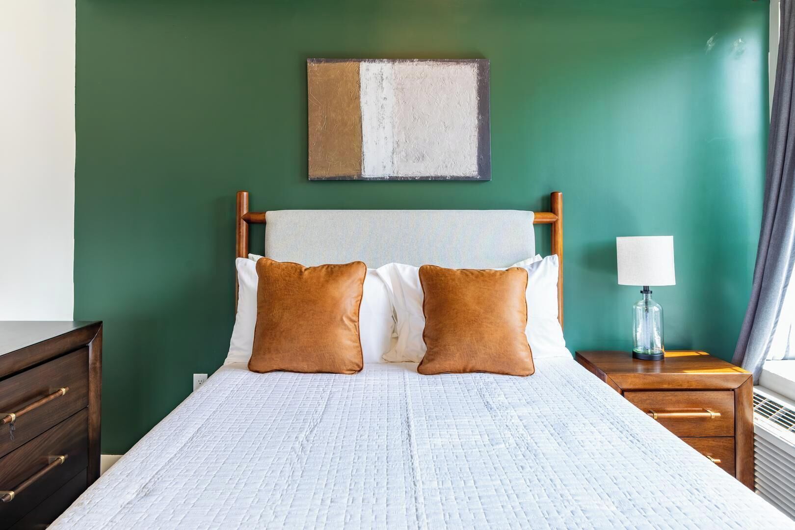Een bed met een witte deken, twee bruine kussens, een houten hoofdbord, een nachtkastje met een lamp en een schilderij aan de muur.