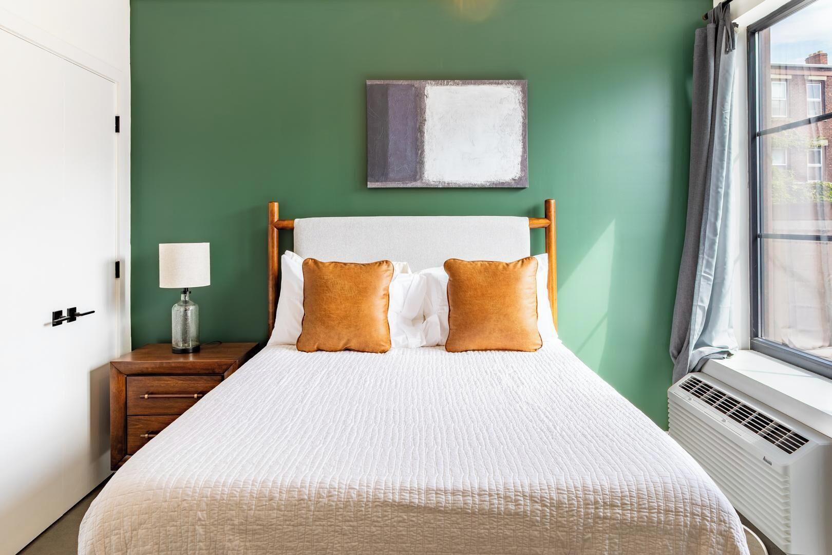 Une chambre d’hôtel avec un lit, une table de chevet, une lampe, un tableau et une fenêtre avec des rideaux.