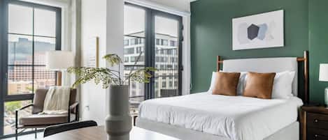 Een moderne hotelkamer met een groot bed, een bureau, een stoel, een vaas met een plant en uitzicht op gebouwen door grote ramen.