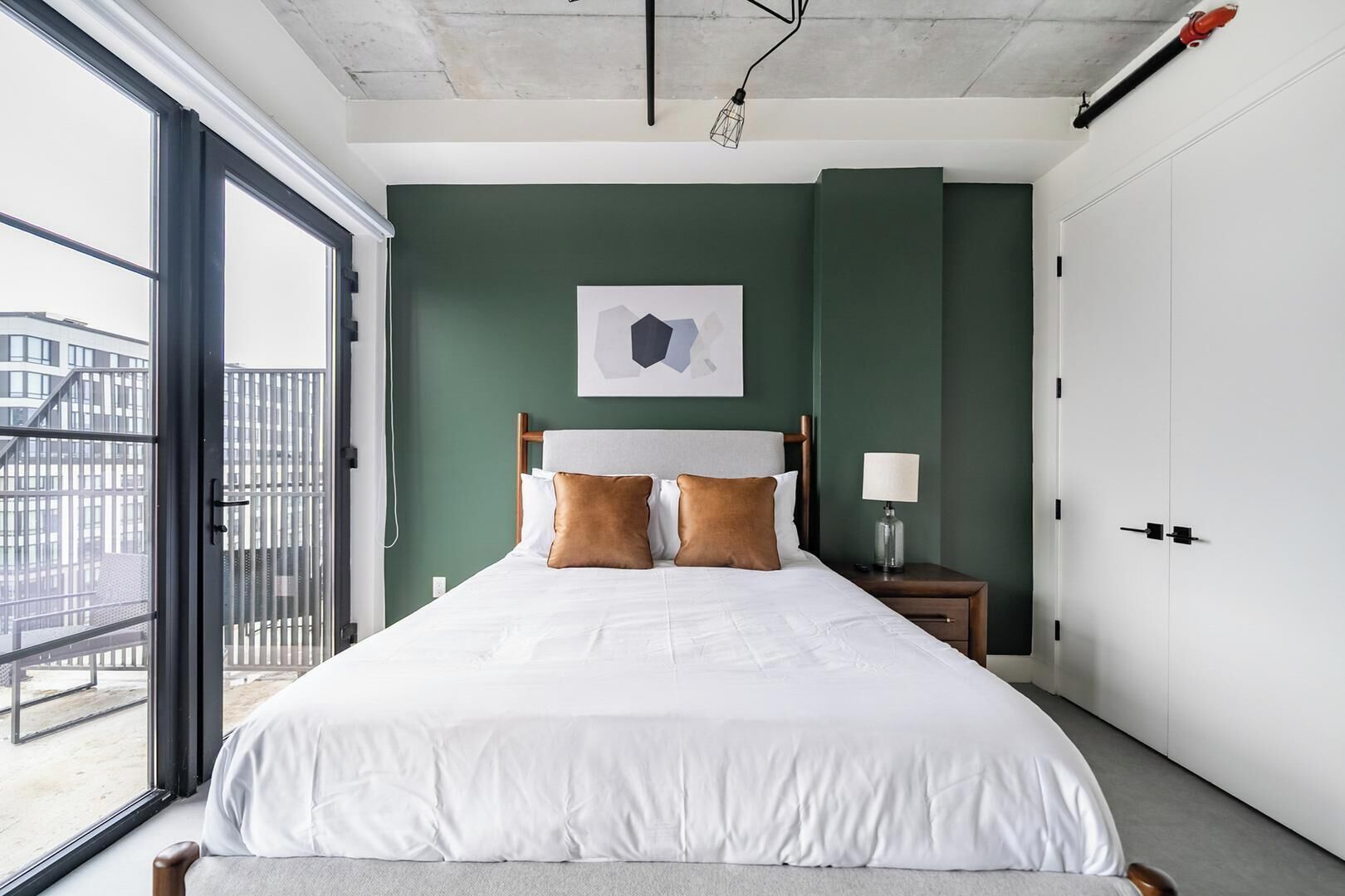 Een slaapkamer met een bed, een nachtkastje, een lamp, een ingelijst beeld en uitzicht door een raam.