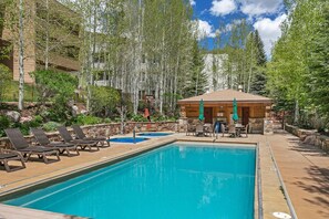 A pool area with lounge chairs, a covered seating area, and a stone wall. - Sun Vail 11b, 3BD Condo (Vail)