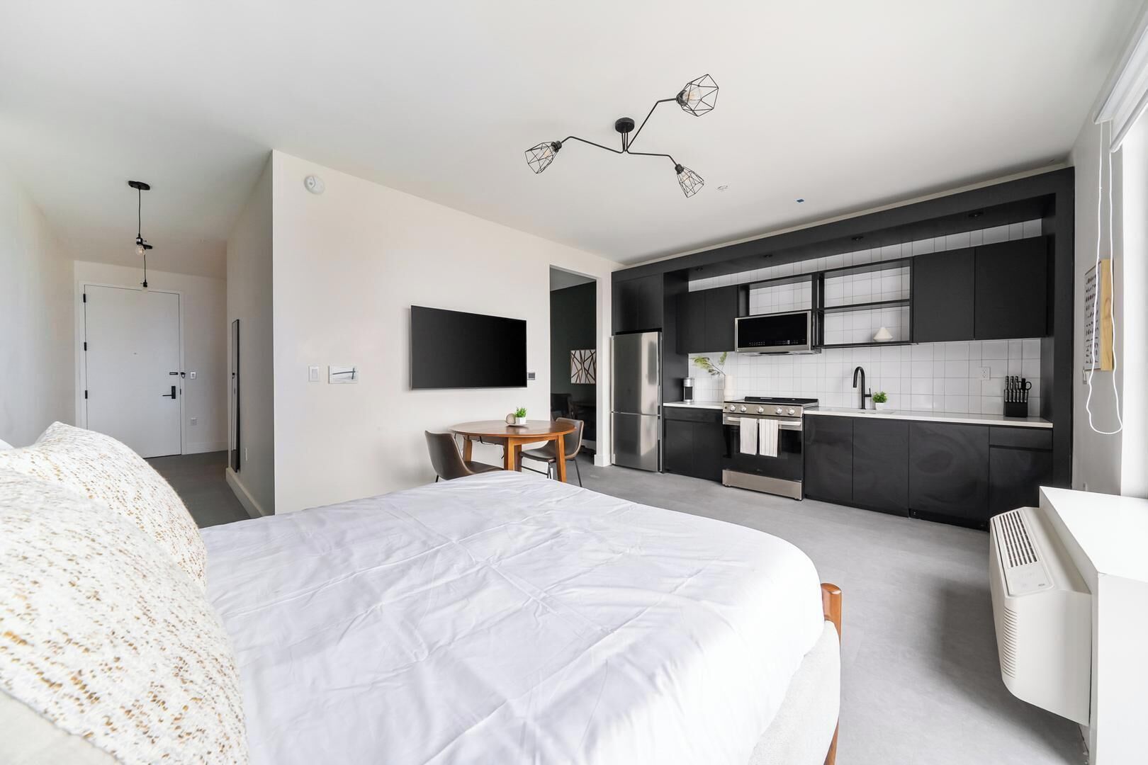 A modern hotel room with a large bed, a dining table, a kitchenette, and a TV mounted on the wall.
