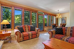 A spacious room with large windows, a wooden sofa, a wooden coffee table, and a chandelier. - Sun Vail 22b, 2BD Condo With Vail Mountain Views (Vail)