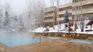 A heated outdoor pool with stone tiles, surrounded by snow-covered trees and a multi-story building. - Sun Vail 22b, 2BD Condo With Vail Mountain Views (Vail)