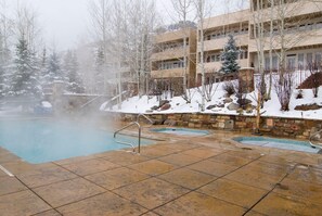 A heated outdoor pool with stone tiles, surrounded by snow-covered trees and a multi-story building. - Sun Vail 22b, 2BD Condo With Vail Mountain Views (Vail)