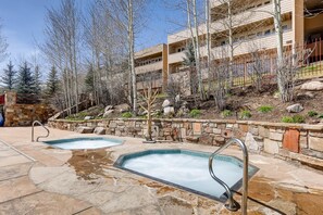 A pool area with two hot tubs, stone walls, and a building with multiple balconies in the background. - Sun Vail 22b, 2BD Condo With Vail Mountain Views (Vail)