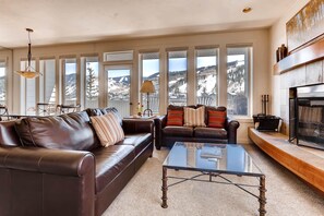 A living room with two leather sofas, a glass coffee table, and a fireplace. Large windows offer a view of snow-covered mountains. - Sun Vail 34b, 3BD Condo With Vail Mountain Views (Vail)