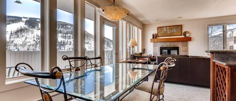 A dining area with a glass table and metal chairs, large windows with a snowy view, a fireplace, and a kitchen area.