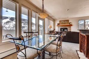 A dining area with a glass table and metal chairs, large windows with a snowy view, a fireplace, and a kitchen area. - Sun Vail 34b, 3BD Condo With Vail Mountain Views (Vail)