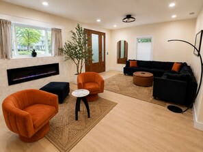 Ein modernes Wohnzimmer mit Kamin, orangefarbenen Sesseln, einer schwarzen Couch und einem großen Fenster mit Blick auf Grünflächen.