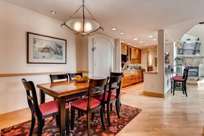 A dining area with a wooden table and chairs, a chandelier, a framed picture on the wall, and a kitchen in the background. - Tyrolean 5, 3BD Condo in Vail Village (Vail)