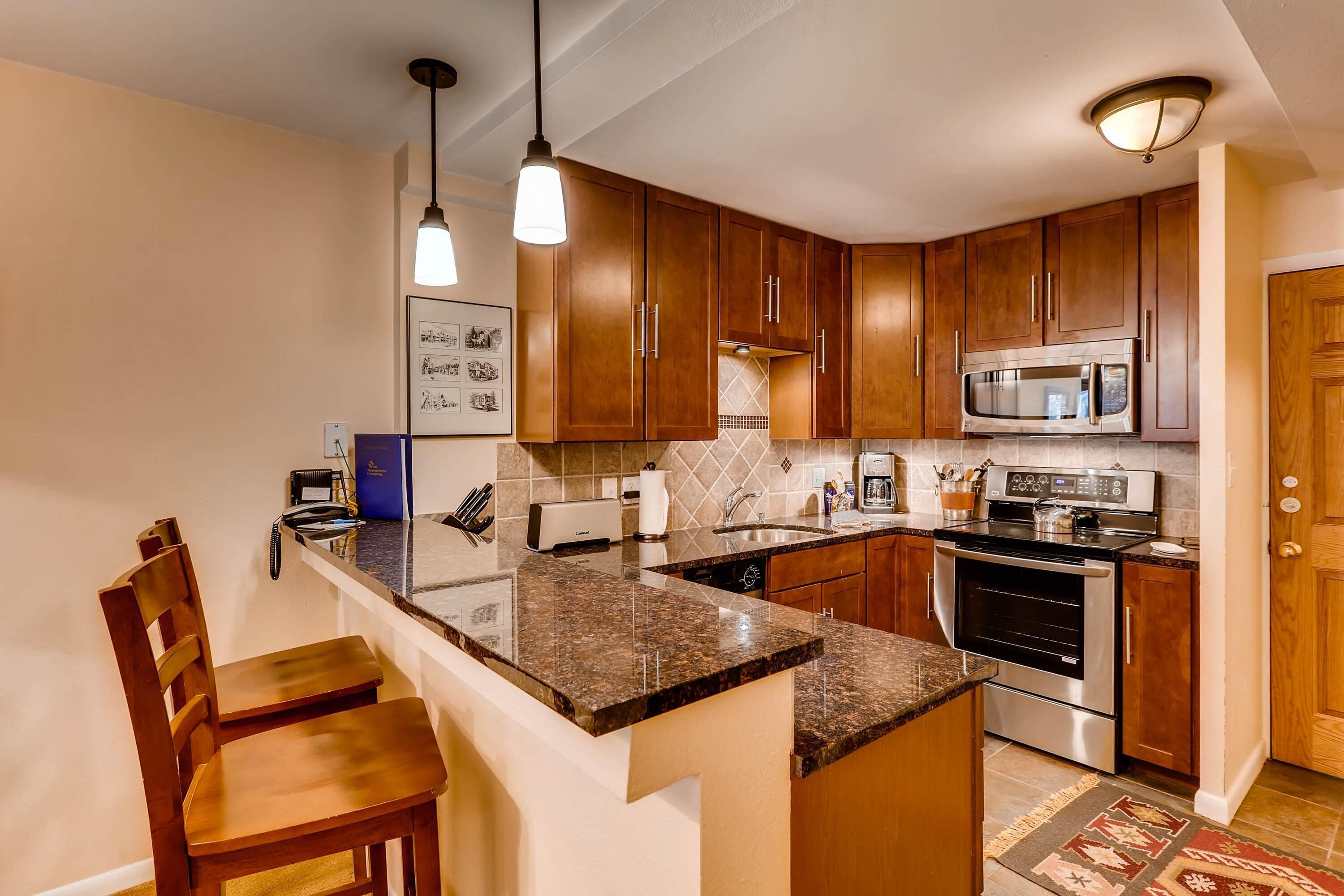 A kitchen with wooden cabinets, a granite countertop, a microwave, an oven, and a dining area with a chair.