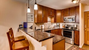 A kitchen with wooden cabinets, a granite countertop, a microwave, an oven, and a dining area with a chair. - Vail Trails East 5B, 1BD Condo on East Side of Vail Village (Vail)