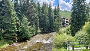 A river flowing through a forest with a building in the background. - Vail Trails East 5B, 1BD Condo on East Side of Vail Village (Vail)