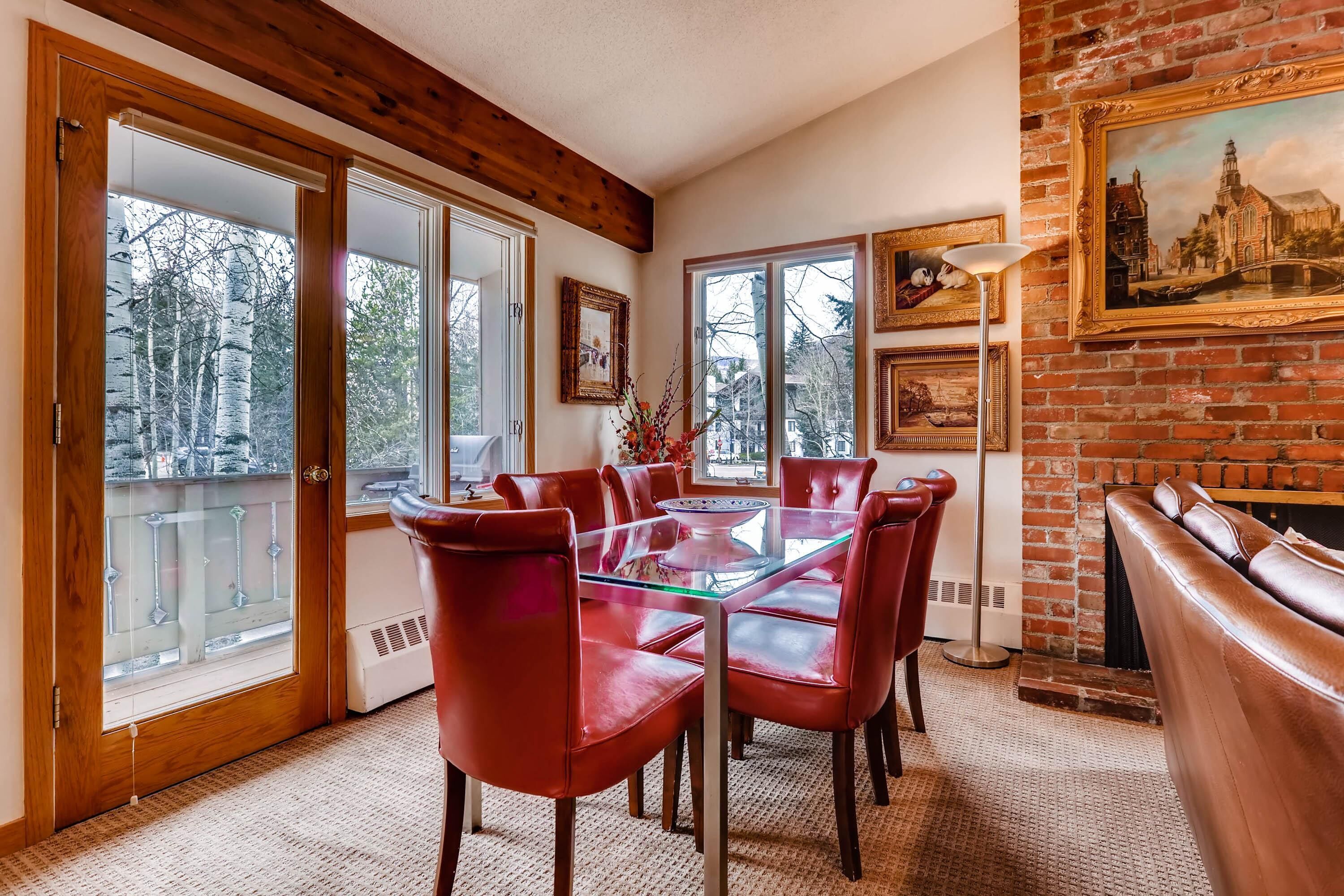 Un coin repas avec une table en verre et des chaises rouges, une cheminée en briques et une vue sur les arbres à travers de grandes portes vitrées.