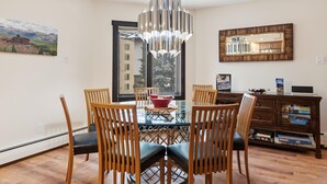 Uma sala de jantar com mesa de vidro, cadeiras de madeira, luminária pendente, espelho e vista para um edifício.