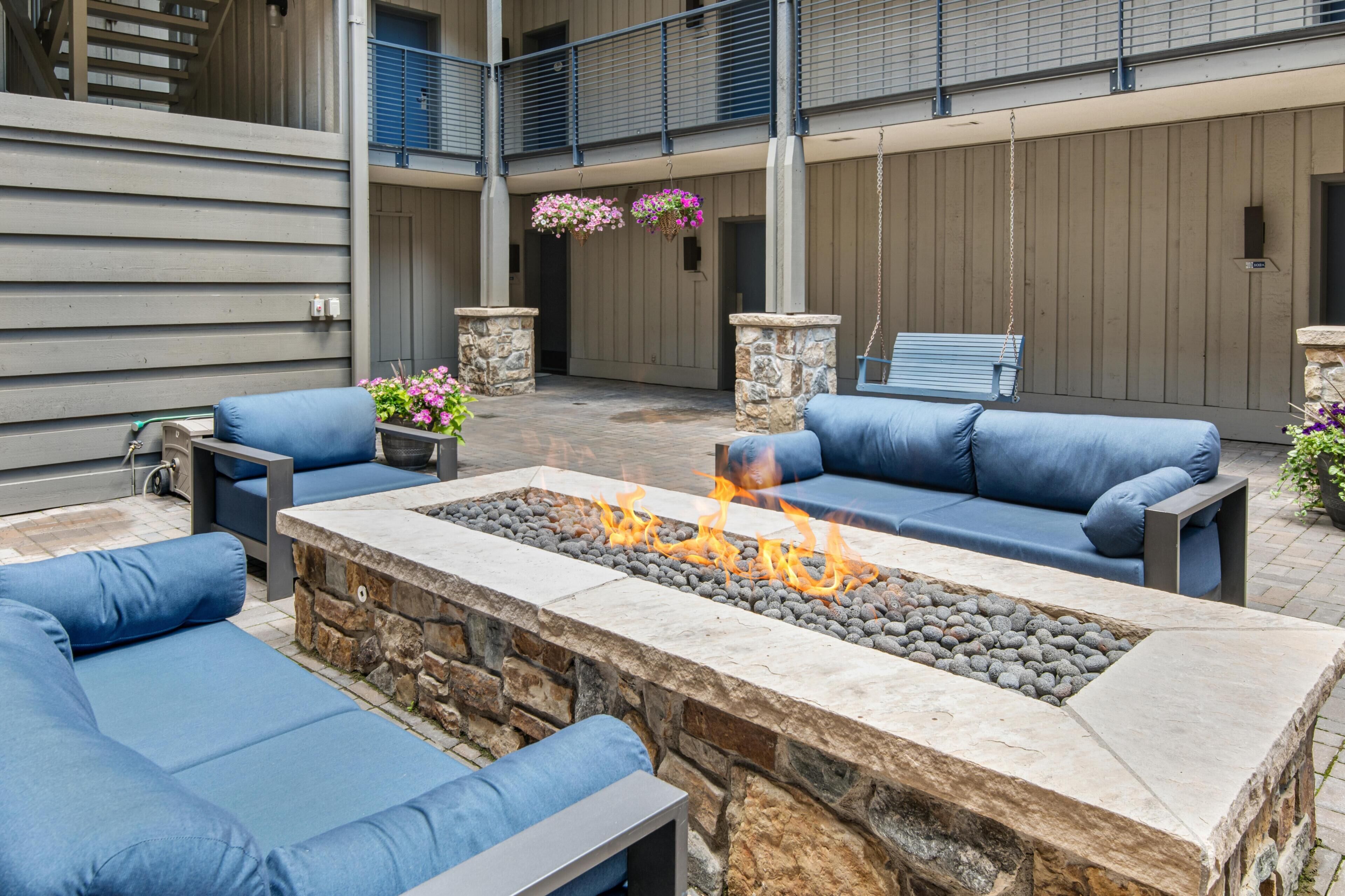 An outdoor seating area with blue cushioned sofas, a stone fire pit with flames, and hanging flower baskets.
