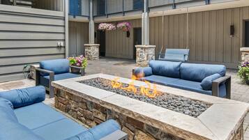 An outdoor seating area with blue cushioned sofas, a stone fire pit with flames, and hanging flower baskets.
