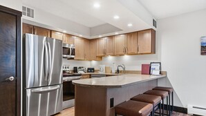 Uma cozinha moderna com geladeira de aço inoxidável, armários de madeira, uma ilha central com banquetas e um micro-ondas acima do balcão.