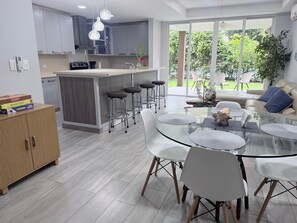 A modern kitchen with a central island, bar stools, a glass dining table with white chairs, and a view of the outdoors. - Apartment in Playa Herradura (Herradura)