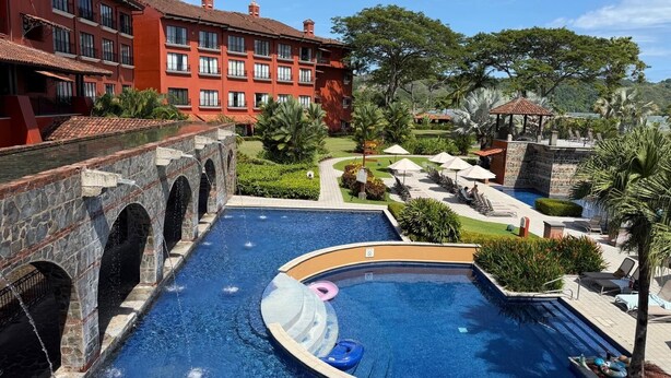 A swimming pool with a stone bridge, lounge chairs, and a red building in the background. - Apartment in Playa Herradura (Herradura)