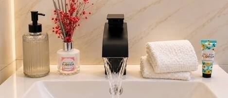 A bathroom sink with running water, a hand soap dispenser, a bottle of hand sanitizer, a vase with dried flowers, a black soap dispenser, and a folded towel.