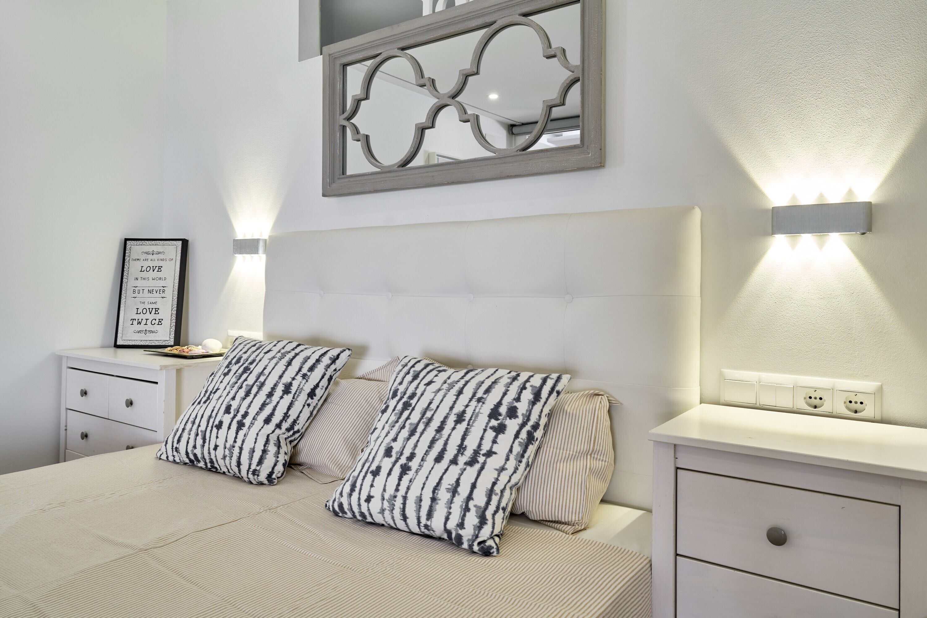 Un dormitorio con cama, dos almohadas, una mesita de noche, una lámpara de pared y un espejo decorativo.