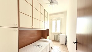 Una habitación de hotel pequeña y sencilla, con cama, armario, ventilador de techo, radiador y ventana.