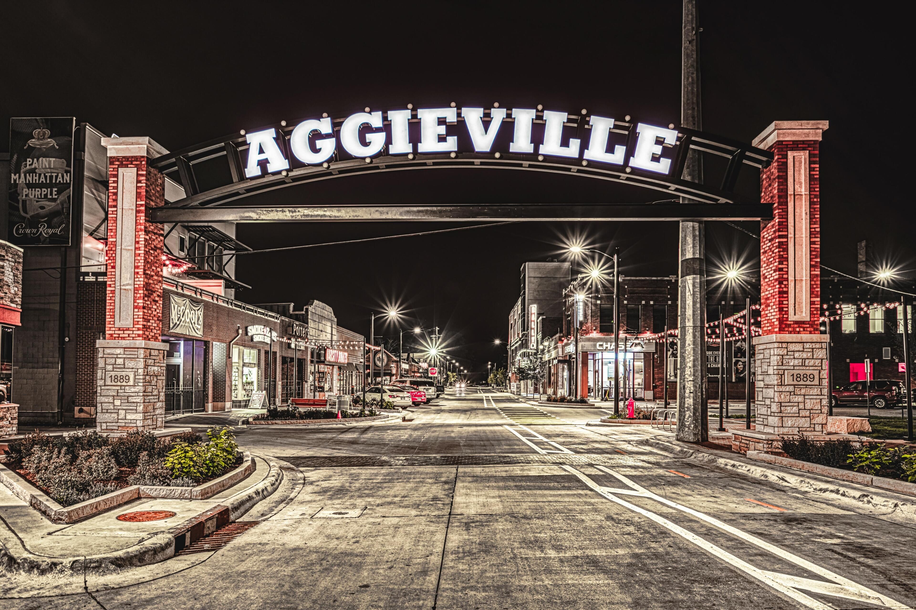 Une vue de rue d’Aggieville, avec une arche imposante sur laquelle est inscrit le nom de la ville, des devantures de magasins et un bâtiment en briques datant de 1889.