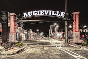 A street view of Aggieville with a prominent archway displaying the town's name, storefronts, and a brick building with the year 1889.