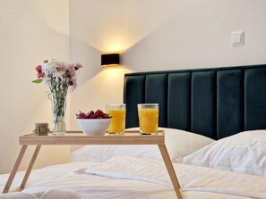 Ein Hotelzimmer mit einem Bett, einem Nachttisch mit einer Schale Obst und zwei Gläsern Orangensaft, einer Vase mit Blumen und einer Wandleuchte.